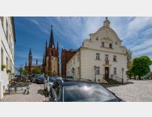Straßenszene mit einem historischen Gebäude rechts im Vordergrund, das eine verzierte Fassade und ein geschwungenes Giebel­design aufweist. Im Hintergrund ist eine rote Backsteinkirche mit hohen, spitzen Türmen und einer Uhr sichtbar, begleitet von geparkten Autos auf dem gepflasterten Straßenabschnitt.