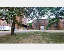 Backsteingebäude eines ehemaligen Industriekomplexes mit großen Fenstern und begrünten Fassaden, umgeben von Bäumen und einer Wiese. Links stehen parkende Anhänger auf einem gepflasterten Hof.