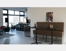 Ein moderner, lichtdurchfluteter Raum mit großen Fenstern, braunen gepolsterten Sitzbänken und schwarzen Tischen. Im Hintergrund ist eine Bar zu sehen, an der eine Person arbeitet, mit Getränken und Lebensmitteln auf der Theke.