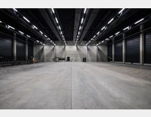 Große, leere Industriehalle mit glattem Betonboden, hohen Decken und einer Reihe von leuchtenden Deckenleuchten. Die Wände bestehen aus grauen Betonflächen, und an den Seiten befinden sich geschlossene, schwarze Rolltore.