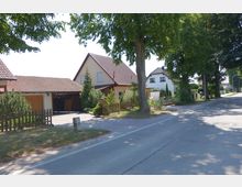 Eine ruhige Wohnstraße mit Einfamilienhäusern, die rote Ziegeldächer und helle Fassaden haben. Der Bereich ist von hohen Bäumen gesäumt, mit kleinen Vorgärten und Holzzäunen vor den Häusern.