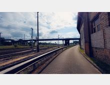 Ein Bahnhofsgelände mit mehreren Gleisen, einer Fußgängerbrücke im Hintergrund und einem gepflasterten Weg neben einem Backsteingebäude mit teils verbarrikadierten Fenstern. Im Hintergrund sind Strommasten und bewachsene Flächen zu sehen, darüber ein bewölkter Himmel.