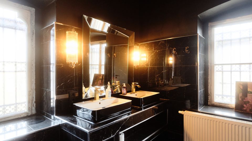 Modernes Badezimmer mit schwarzen, marmorierten Wand- und Bodenfliesen, Doppelwaschbecken und großem Spiegel mit warmen Wandleuchten. Zwei große Fenster lassen Tageslicht herein; darunter befinden sich eine breite Fensterbank und ein Heizkörper.