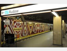 U-Bahn-Station mit der Beschilderung „U7 Rudow“. Die Wand der Station ist mit bunten geometrischen Mustern in Rot-, Gelb- und Weißtönen gestaltet, die Rädern und Maschinen ähneln.