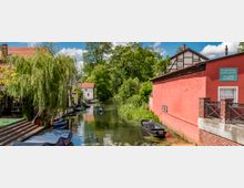 Kanallandschaft in einer kleinstädtischen Umgebung mit kleinen Booten, einem roten Backsteingebäude rechts und grünem Laub entlang des Wassers. Im Hintergrund sind Fachwerkhäuser und Bäume zu sehen, während eine Person in einem Kajak auf dem Wasser paddelt.