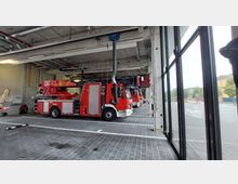 Mehrere rote Feuerwehrfahrzeuge stehen in einer modernen Fahrzeughalle mit grauem Boden und großer Fensterfront. Die Halle ist mit Lüftungs- und Schlauchsystemen ausgestattet, und im Hintergrund sind weitere Fahrzeuge und technische Leitungen zu sehen.