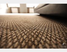 Nahaufnahme eines grob texturierten Teppichbodens in Braun- und Beigetönen, der den Vordergrund dominiert. Im Hintergrund sind unscharfe, helle Fenster sowie die Kante eines Sofas zu sehen.