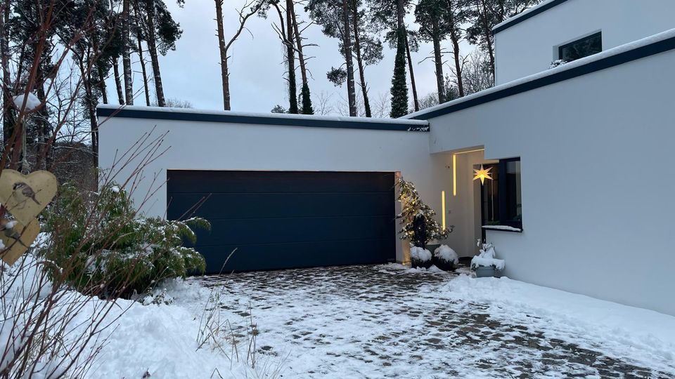 Moderner, weißer Flachdach-Bungalow mit großer dunkler Garagentür und gepflasterter, teilweise verschneiter Einfahrt. Im Hintergrund stehen hohe Kiefern, am Hauseingang ist eine beleuchtete Stern-Dekoration zu sehen.