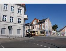 Straßenecke in einer Kleinstadt mit mehrstöckigen Gebäuden im historischen Stil, darunter ein weißes Gebäude links und ein rot-weißes Fachwerkhaus mit Geschäften im Erdgeschoss rechts. Auf der Straße wartet eine Pferdekutsche an einer Ampel.