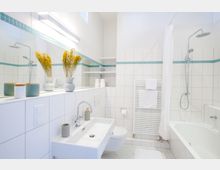 Helles Badezimmer mit weißen Fliesen und einer türkisfarbenen Bordüre, ausgestattet mit einer Badewanne, einer Regendusche, einem Waschbecken und einem wandhängenden WC. Dekoriert mit gelben Trockenblumen in Vasen, einem großen Spiegel und dunklen sowie weißen Accessoires.