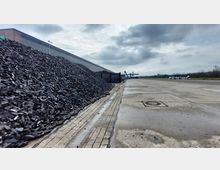 Eine große Lagerfläche mit gepflasterter und betonierter Oberfläche, links ein großer Haufen dunkelgrauer Kohle oder Schlacke. Im Hintergrund befinden sich Kräne und ein Lagergebäude, während rechts eine offene, leere Fläche sowie einige Bäume und Strukturen sichtbar sind. Der Himmel ist bewölkt.