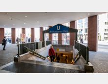 Eingang zur U-Bahn-Station "Bhf Potsdamer Platz" mit einer Treppe, die nach unten führt. Die Umgebung zeigt einen überdachten Bereich mit roten Backsteinpfeilern und Fußgängern im Hintergrund.