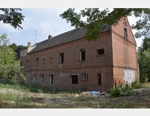 Backstein-Gebäude mit rechteckigen Fenstern, teils ohne Glas, umgeben von hohem Gras und Bäumen. Das Gebäude wirkt verlassen, mit Trümmern und Müll im Vordergrund.