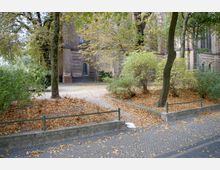 Gartenansicht mit Herbstlaub, umgeben von Bäumen und Büschen, im Vordergrund ein gepflasterter Weg mit niedriger Abgrenzung; im Hintergrund eine Backsteinkirche mit gotischen Fenstern.