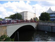 Straßenbrücke mit roten Geländern über einen Fluss, im Hintergrund ein mehrstöckiges helles Gebäude und ein modernes Gebäude mit einem spitz zulaufenden Dach. Autos und Fußgänger befinden sich auf der Brücke, daneben Bäume und Gehwege.