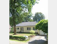 Einfamilienhaus im Bungalowstil mit hellem Putz und dunklem Ziegeldach, davor ein kleiner Vorgarten mit Rasen, Sträuchern, Steinen und großen Bäumen. Rechts führt eine gepflasterte Einfahrt zur angebauten Garage.