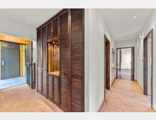 Heller Hausflur mit beigefarbenen Bodenfliesen, weißen Wänden und Einbauschrank aus dunklem Holz mit Lamellentüren und Nische. Der Gang führt zu mehreren Türen mit Holzrahmen; links ist der Eingangsbereich mit großer Holztür und seitlichem Fenster zu sehen.