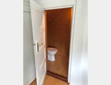 Offene Tür zu einer kleinen Toilette mit holzvertäfelten Wänden und einem Holzboden. Die Tür ist weiß und befindet sich in einem Raum mit heller Wand und einem Laminatboden.