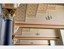 Innenraum mit dekorativer Decke und Säulen. Die Decke ist mit geometrischen und floralen Mustern verziert, umgeben von farbigen Zierleisten und rechteckigen Balken; mehrere klassische Kronleuchter hängen von der Decke.