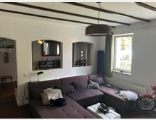 Wohnzimmer mit einer großen L-förmigen Couch in dunklem Farbton, einem kleinen weißen Couchtisch und einem Motorradreifen, der als Dekoration dient. Im Hintergrund befinden sich eine Wand mit drei Durchgängen, die Blick auf einen Flur und eine weitere Einrichtung ermöglichen. Ein Fenster mit grünen Vorhängen lässt Tageslicht herein.