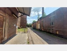 Industriegelände mit Backsteingebäuden, die einen schmalen, gepflasterten Hof begrenzen. Links ein überdachter Bereich, rechts ein Gebäude mit großen Fenstern, teilweise beschädigt; im Hintergrund sind ein Schornstein und Bäume zu sehen.