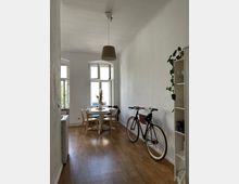 Schmaler Wohnraum mit hellem Holzfußboden, einem runden Esstisch mit vier Stühlen vor einem großen Fenster, einem schwarzen Fahrrad an der Wand sowie einem Regal mit Geschirr und einer Hängepflanze. Eine einfache Hängelampe hängt von der Decke.