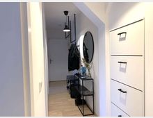 Ein schmaler Flur mit hellem Laminatboden, weißen Wänden und moderner Einrichtung. Auf der rechten Seite befindet sich ein weißer Schuhschrank mit schwarzen Griffen, links ein runder Wandspiegel über einem schmalen Metalltisch mit Dekoration. Im Hintergrund sind eine weiße Tür und eine Garderobe mit Jacken erkennbar.