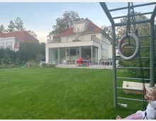 Blick über einen großen Rasen auf ein modernes, weißes Wohnhaus mit großen Glasfronten, Terrasse und Dachterrasse unter rotem Satteldach. Rechts im Vordergrund steht ein Spielgerüst mit Leiter und hängenden Turnringen, umgeben von Bäumen und weiteren Häusern im Hintergrund.