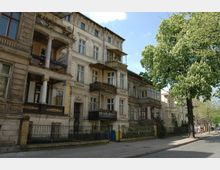 Alt-Text: Straßenansicht von alten Mehrfamilienhäusern im Stil des 19. Jahrhunderts mit verzierten Fassaden, Balkonen und schmiedeeisernen Geländern. Im Vordergrund steht ein großer blühender Baum, und auf dem Gehweg sind Mülltonnen sowie einige Passanten erkennbar.