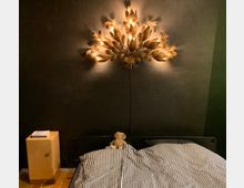 Ein Schlafzimmer mit einer schwarzen Wand, an der eine dekorative Wandleuchte in Blätterform hängt und warmes Licht ausstrahlt. Im Vordergrund steht ein Bett mit schwarz-weiß gestreiftem Bettbezug und einem Stofftier darauf, daneben ein kleiner Holztisch mit Büchern.