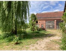 Ein Bauernhof mit einem großen, roten Backsteingebäude und einer alten Holztür, umgeben von einer grünen Wiese. Im Vordergrund steht eine Weide mit überhängenden Zweigen, und auf der Wiese liegen Holzstapel sowie mehrere Blumentöpfe.