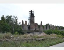 Industriegelände mit stillgelegten Schornsteinen und einem zentralen, von Gerüsten umgebenen großen Kamin, umgeben von überwucherter Vegetation und teils verwitterten Strukturen. Im Vordergrund sind Gras, Büsche und Erdhügel sichtbar.