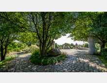 Gepflegter Parkbereich mit gepflastertem Rundweg, umgeben von Bäumen und dichter Vegetation. Im Hintergrund Sitzbänke und Skulpturen, auf denen oder um die sich Personen aufhalten, mit Blick auf eine grüne Landschaft.