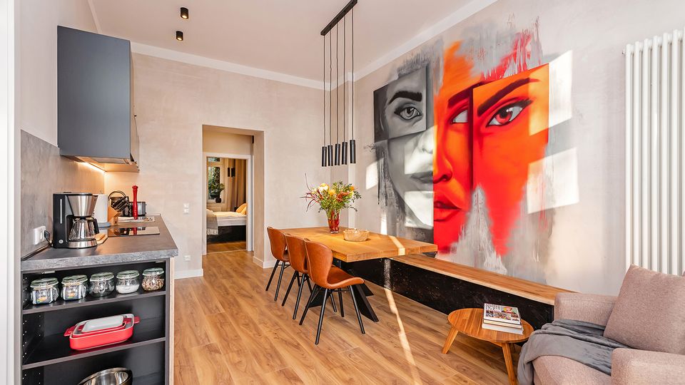 Offener Wohn- und Essbereich mit integrierter Küchenzeile, Holzfußboden und modernem Esstisch mit Stühlen unter schlanken Pendelleuchten. Auffällig ist ein großflächiges Wandgemälde mit einem Gesicht in Grau- und Rottönen, daneben ein Sofa und ein weißer Heizkörper.