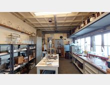 Innenansicht eines historischen Chemielabors mit Laborgeräten, Regalen voller Glaswaren und Chemikalien, einem zentralen Arbeitstisch und einer Werkbank entlang der Fenster. Die Wände und Möbel wirken alt, mit sichtbaren Gebrauchsspuren.