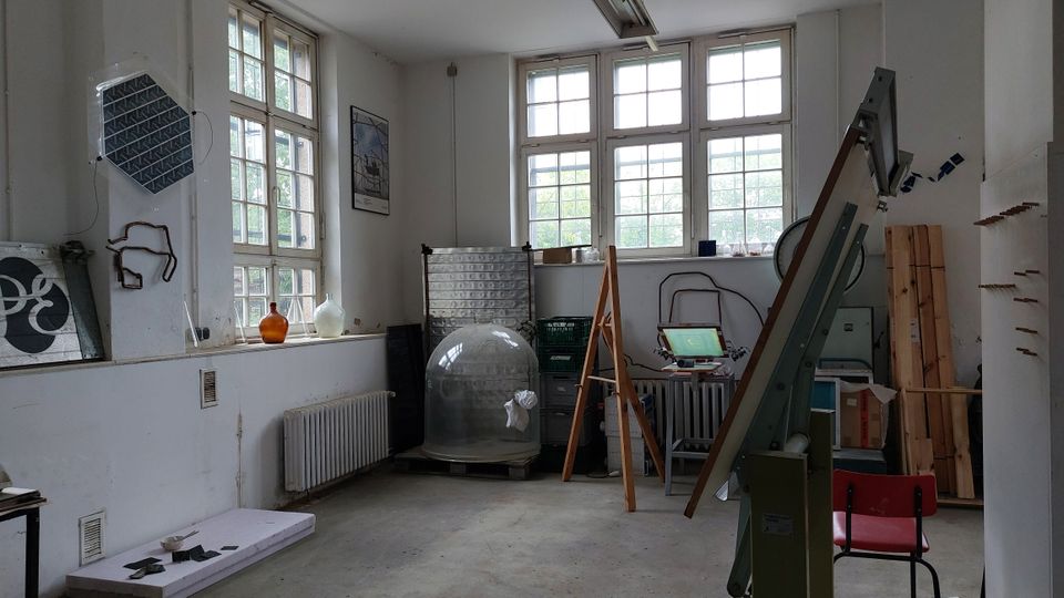 Ein Werkraum mit weißen Wänden, großen Fenstern mit Holzrahmen und verstreuten Gegenständen wie einer Glaskuppel, Geräten, Werkzeugen und Materialien. Im Vordergrund stehen ein Holzgestell und eine rote Stuhl.