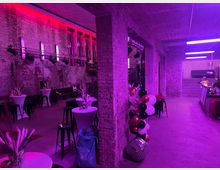 Ein Veranstaltungsraum mit Ziegelwänden, lila und pink beleuchtet. Der Raum verfügt über Stehtische mit Dekoration, Barhocker und eine seitlich gelegene Theke; an einer Wand sind bunte Luftballons befestigt.