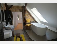 Modernes Badezimmer in einem Dachgeschoss mit freistehender Badewanne auf einer erhöhten Plattform, Waschbecken auf einer Holzablage, einer Backsteinwand und schrägem Dach mit großem Dachfenster. Im Hintergrund ist ein Flur mit Holzboden sichtbar.