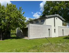 Modernes Einfamilienhaus mit weißer Putzfassade und einer Verkleidung aus horizontalen Holzlatten. Das Gebäude steht auf einem gepflegten grünen Rasen, umgeben von großen Bäumen und blauem Himmel.