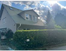 Zweistöckiges Einfamilienhaus mit grauem Putz und grünem Satteldach, umgeben von einer Hecke und einem Zaun. Im Hintergrund sind Nadelbäume und ein bewölkter Himmel mit Sonnenschein zu sehen.