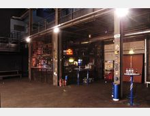 Innenraum eines industriell gestalteten Veranstaltungsortes mit sichtbaren Metallstrukturen, erhöhten Laufstegen und einer Bar, die durch ein Gitter abgetrennt ist. Im Vordergrund stehen hohe Stehtische mit Getränkeflaschen, im Hintergrund leuchten ein "Red Bull"-Neonschild und andere Lichtquellen.