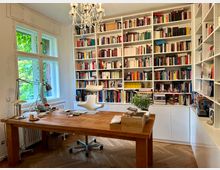 Helles Arbeitszimmer mit einer großen weißen Bücherwand, gefüllt mit Büchern und Dekorationsgegenständen. Im Vordergrund ein massiver Holztisch mit Schreibutensilien, eine beige Bürostuhl und eine Schreibtischlampe; links ein Fenster mit Blick auf grüne Blätter und ein Kronleuchter an der Decke.