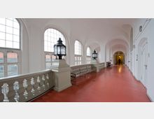 Ein heller Innenflur mit weiß gestrichenen Wänden und hohen Segmentbogenfenstern, die viel Tageslicht hereinlassen. Der rote Boden, eine weiße Balustrade mit stilisierten Laternen und Holzbänke entlang der Balustrade prägen den Raum; im Hintergrund ist eine einzelne Person zu sehen.