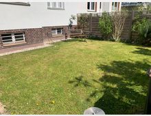 Ein kleiner Garten mit einer Rasenfläche, begrenzt durch einen Holzzaun mit Sträuchern und Pflanzen. Im Hintergrund steht eine Metallbank vor der Hauswand eines weißen Gebäudes mit Klinker-Sockel.