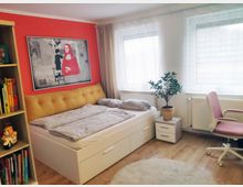 Helles Schlafzimmer mit roter Akzentwand, auf der ein gerahmtes Kunstbild hängt. Ein Bett mit gelbem gepolstertem Kopfteil steht vor der Wand, daneben ein Nachttisch mit einer Topfpflanze und daneben zwei große Fenster mit weißen Vorhängen; links ein Bücherregal und rechts ein rosa Schreibtischstuhl.