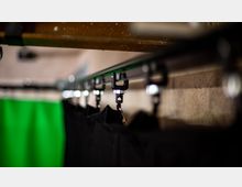 Nahaufnahme einer Vorhangschiene mit mehreren schwarzen Vorhängen, die an silbernen Haken aufgehängt sind. Im Hintergrund ist eine grüne Fläche sowie eine beige Wand unscharf zu erkennen.