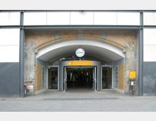 Eingang eines Bahnhofsgebäudes mit einem gemauerten Rundbogen aus roten Ziegeln und einer integrierten Uhr über der Tür. Der moderne Eingang verfügt über automatische Glastüren, darüber hängt ein gelbes Schild mit Hinweistext.