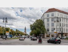 Straßenkreuzung in einer Stadt mit mehreren Fahrzeugen und einem Motorradfahrer, flankiert von einem großen, weißen Eckgebäude mit rotem Dach und Geschäftsräumen im Erdgeschoss. Im Hintergrund sind Bäume und ein hoher Kirchturm erkennbar, während Laternen und Verkehrsschilder die Szene ergänzen.
