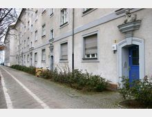 Stadthausfassade mit hellgrauer Putzfassade, blau gestrichenen Türen und weißen Fensterrahmen; über den Türen befinden sich kleine dekorative Skulpturen. Ein bepflanzter Streifen trennt das Gebäude von einem gepflasterten Gehweg mit Radspuren.