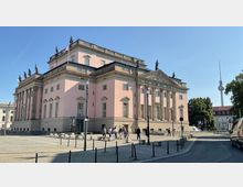 Großes, historisches Gebäude im neoklassizistischen Stil mit rosa Fassade und Säulen, verziert mit Statuen auf dem Dach. Im Hintergrund sind Bäume, ein moderner Fernsehturm und weitere Gebäude zu sehen; im Vordergrund ist ein gepflasterter Platz mit einigen Passanten.